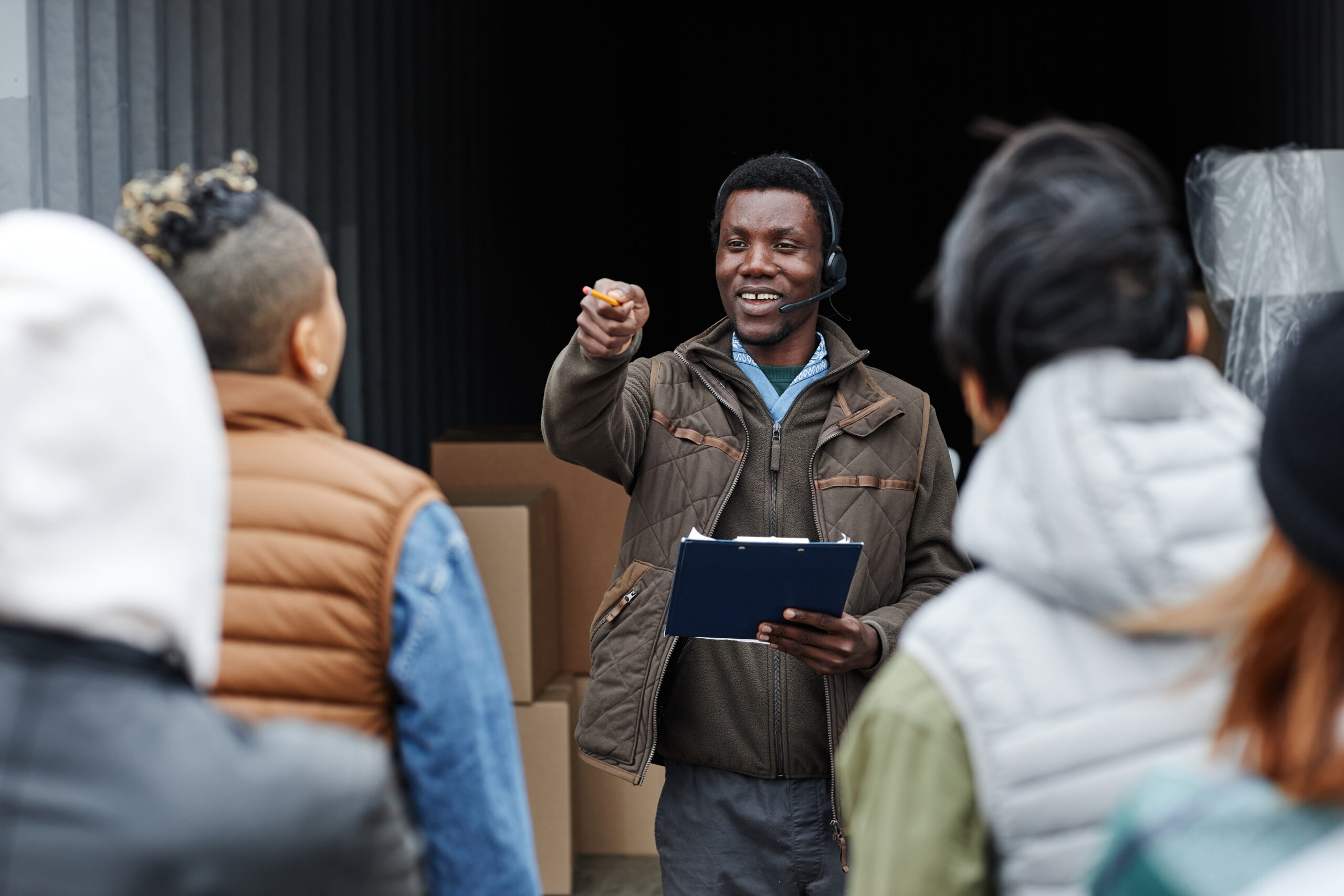 Waist up portrait of smiling auctioneer standing by container at shipping port and wearing headset taking bids from buyers
